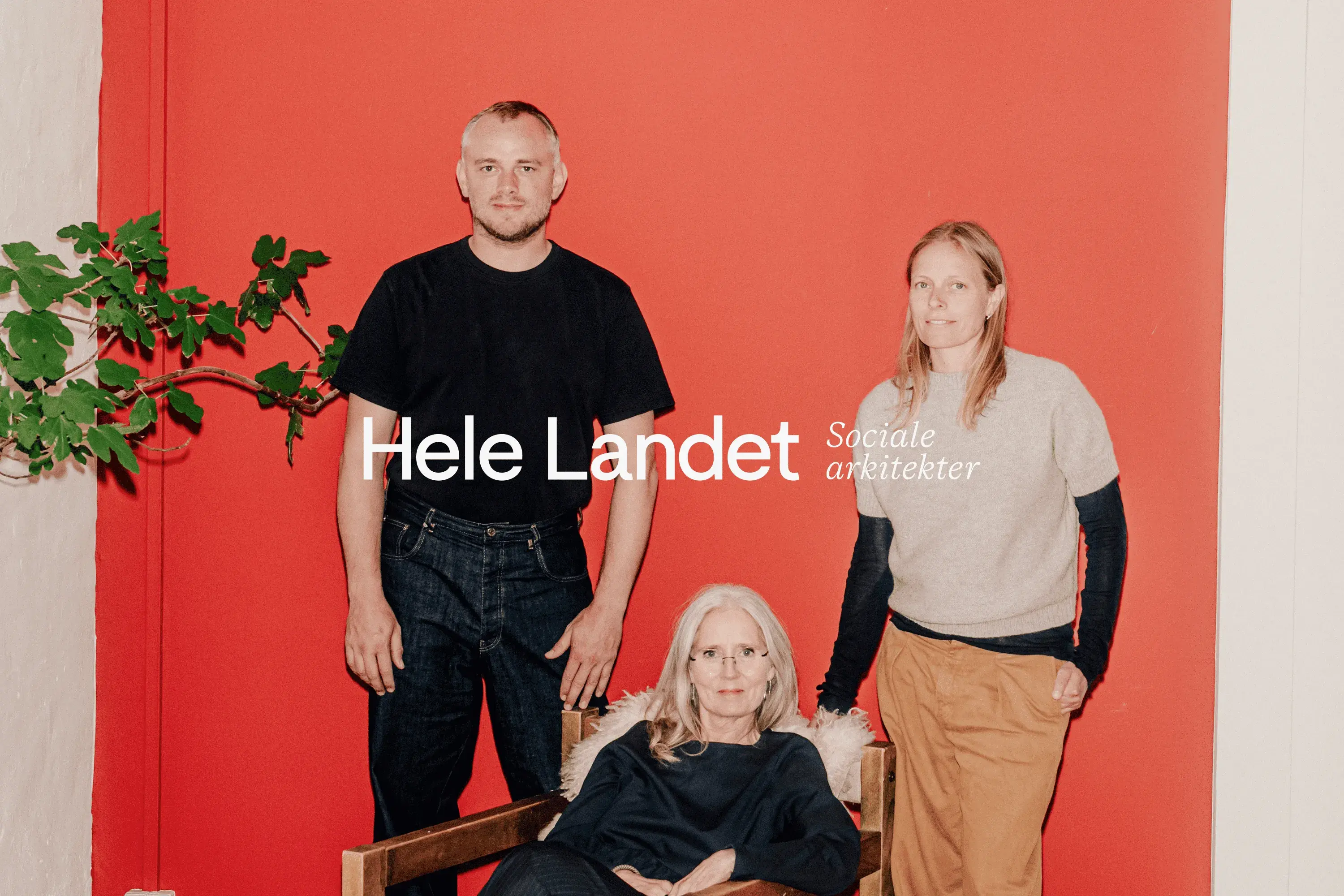 Three adults—one seated, two standing—pose against a red wall with white "Hele Landet" text and a leafy plant at left.