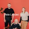 Three adults—one seated, two standing—pose against a red wall with white "Hele Landet" text and a leafy plant at left.