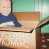 A baby with a pacifier sits in an open cardboard box on a patterned rug, in front of a green wall.