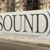 Large white sign with the word "SOUND" in bold letters, surrounded by sound wave graphics, placed on a street with scaffolding behind.