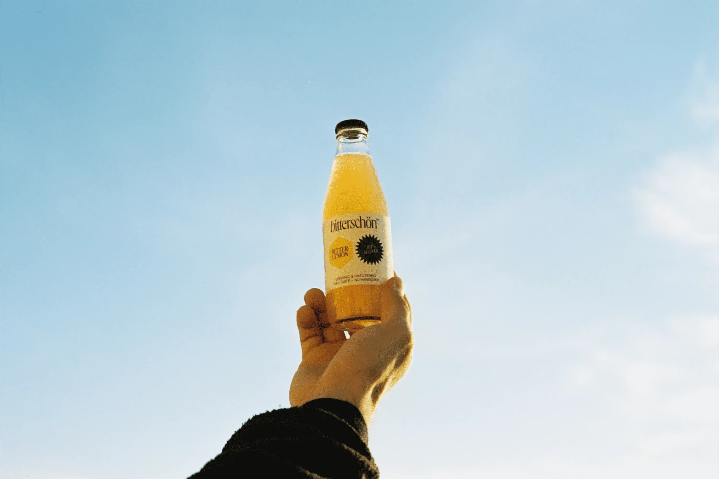 A hand holds up a bottle of orange juice against a clear blue sky, showcasing the label and contents.