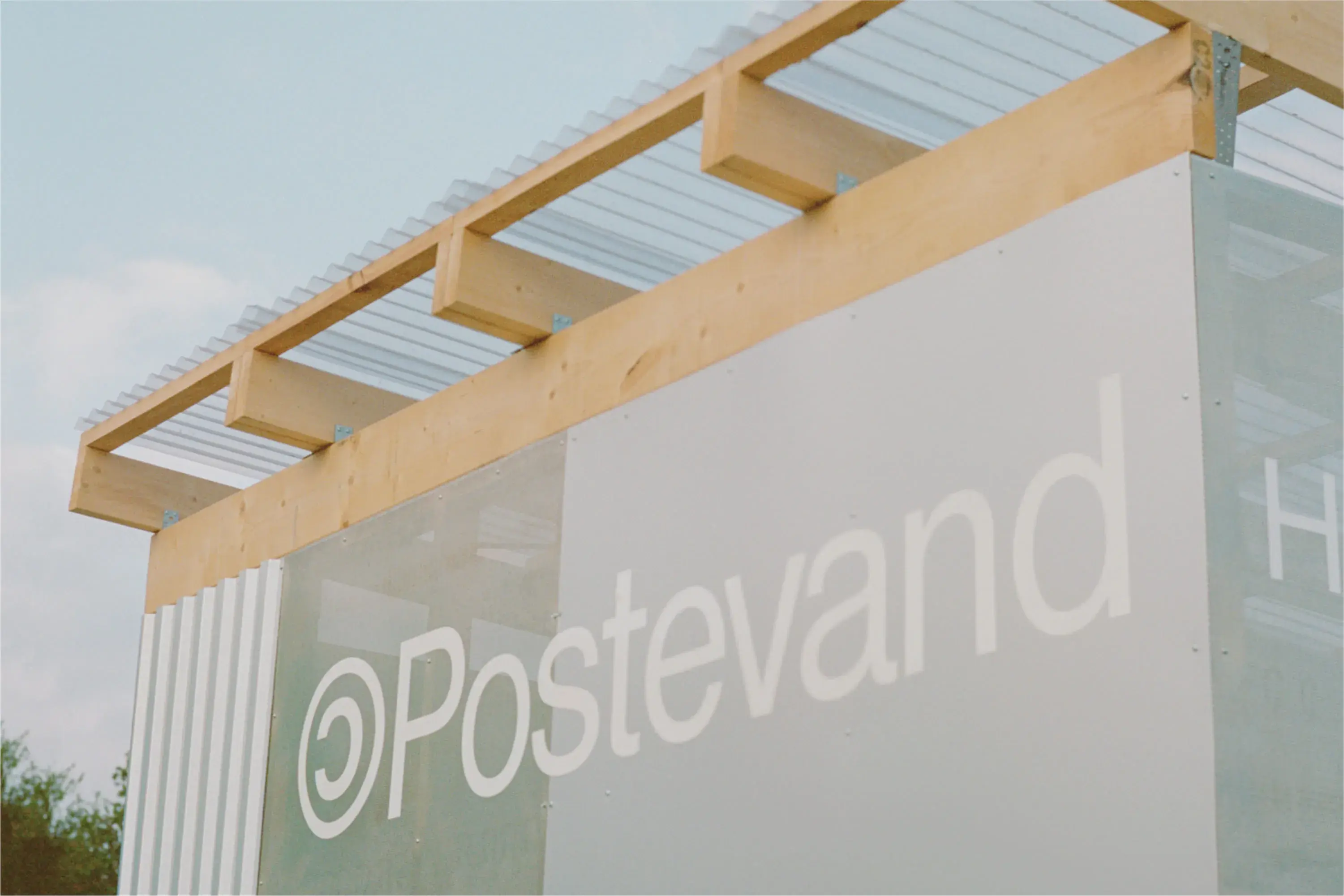 Sign reading "Postevand" on a modern structure with wooden beams and clear panels, set against a blue sky.